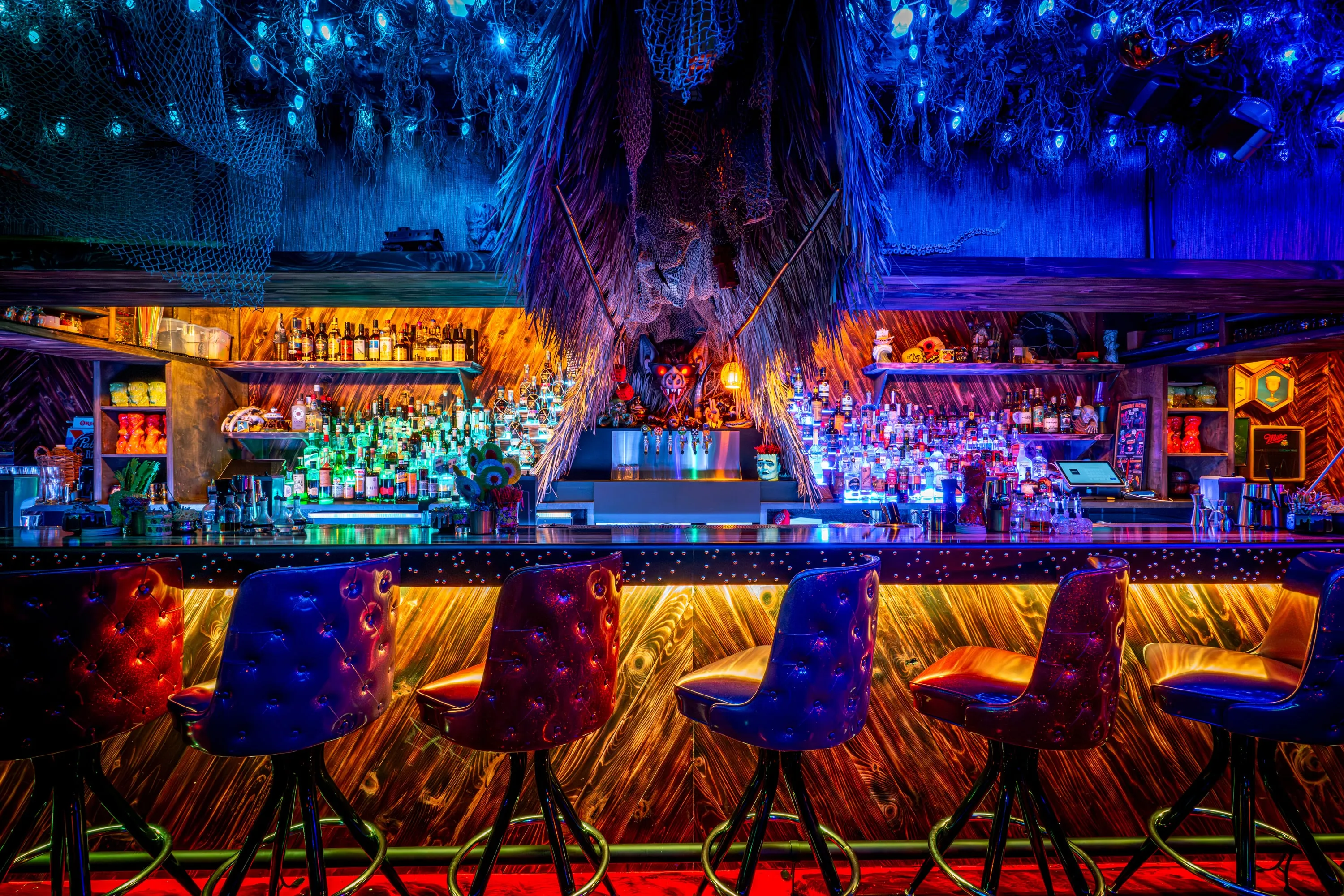 Bar at Paradise Lost under a canopy of blue-lit foliage, with red-lit back bar and tufted stools
