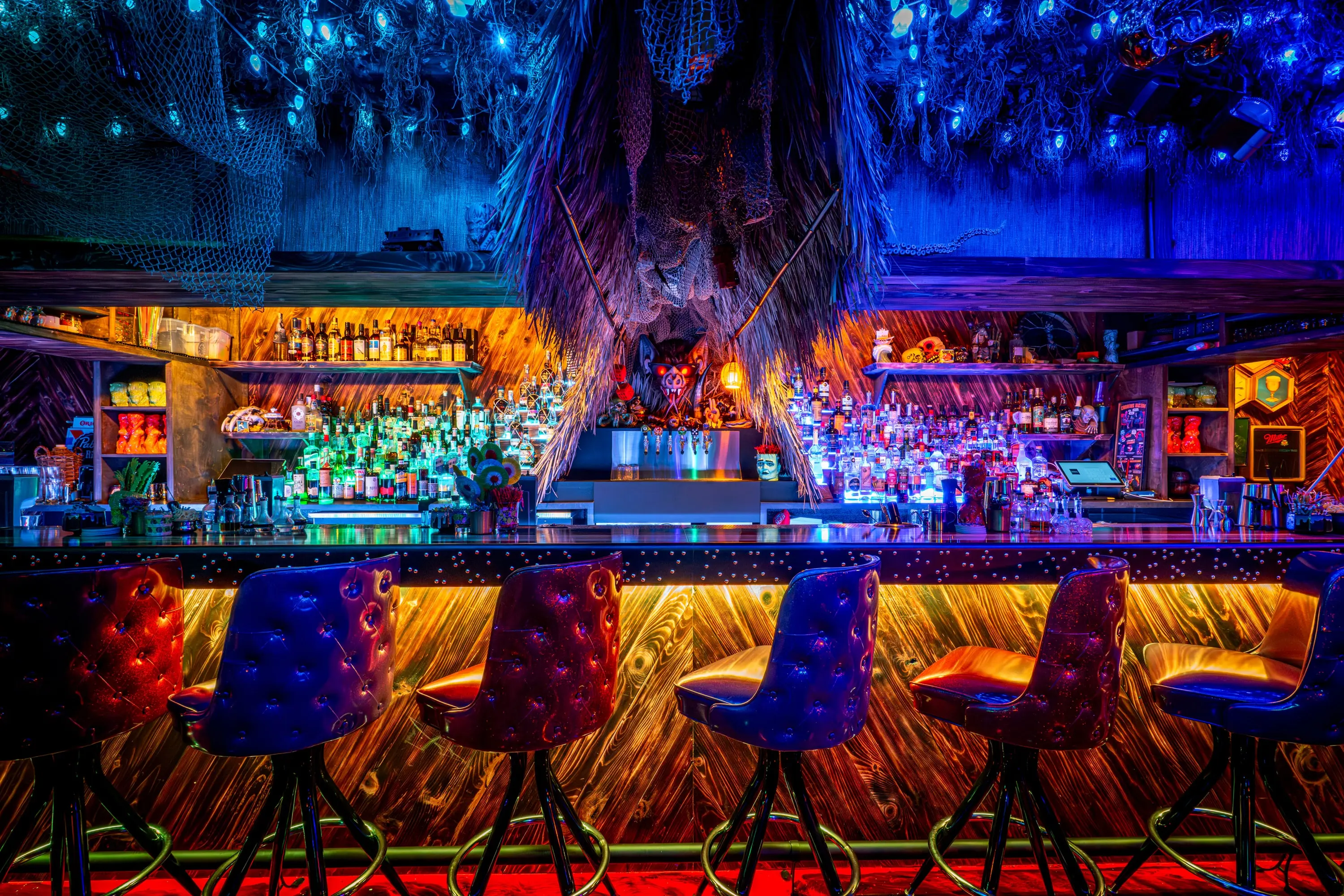Bar at Paradise Lost under a canopy of blue-lit foliage, with red-lit back bar and tufted stools