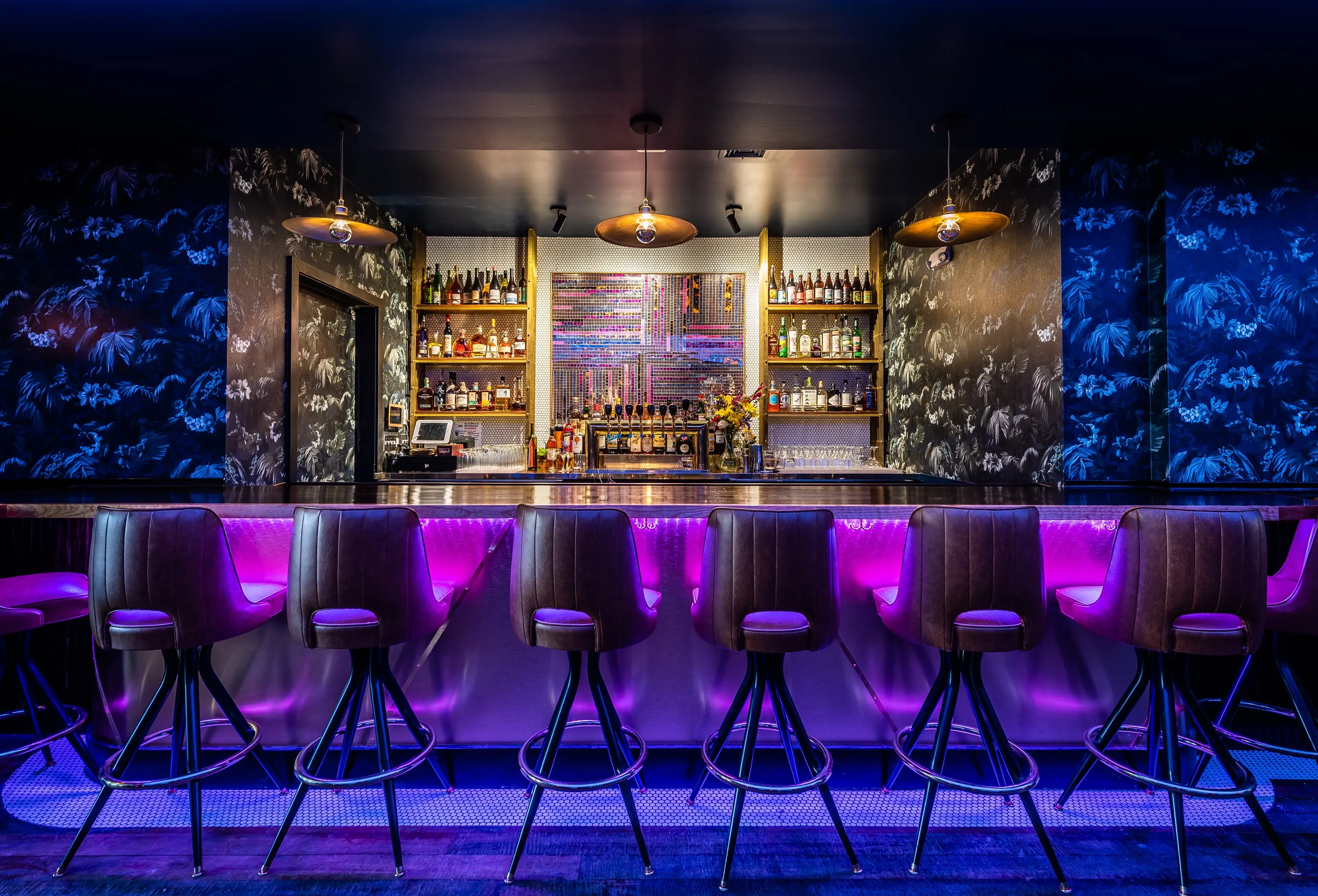 Bar at No Kisses lit violet with dark patterned wallpaper and stools