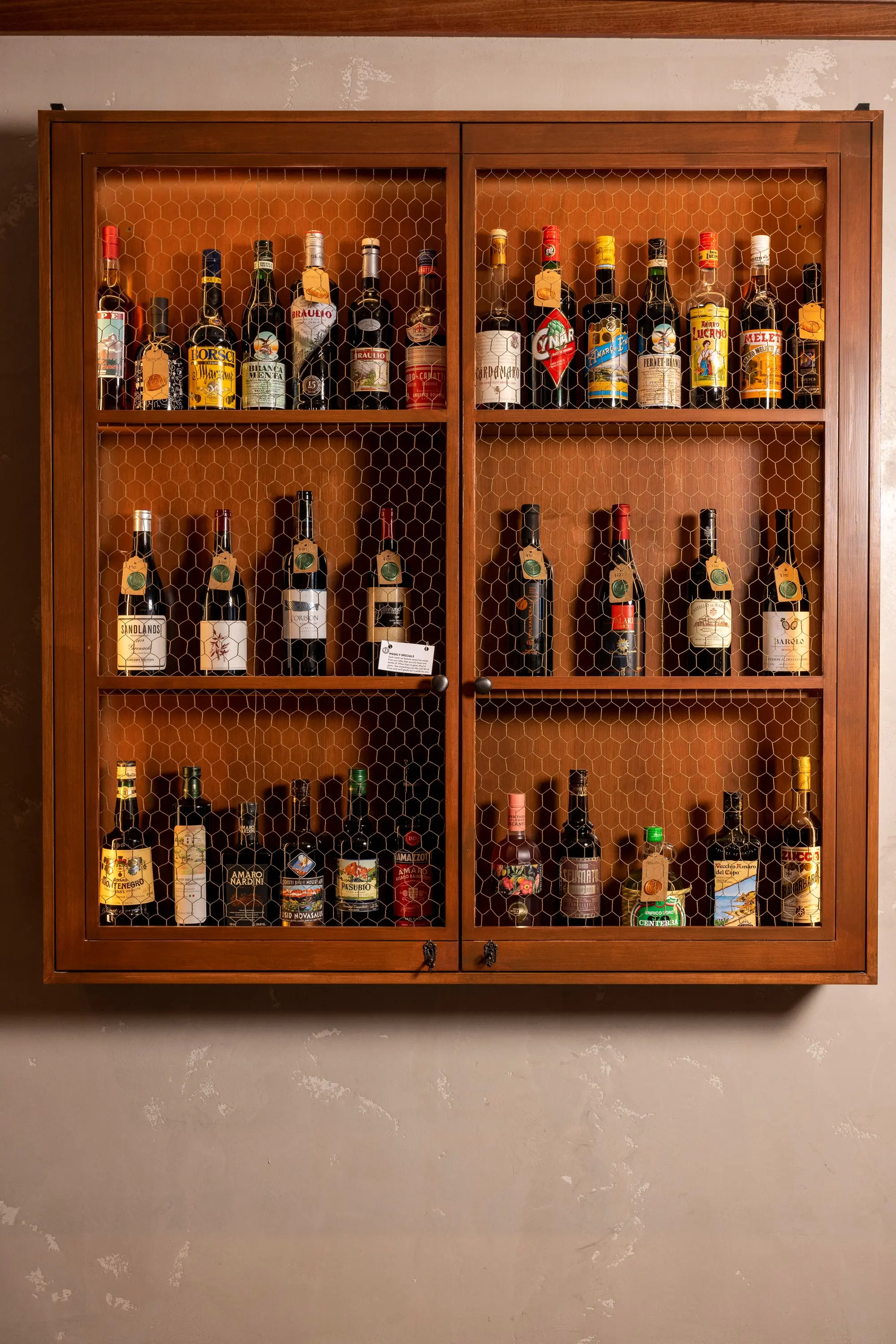 Wood and wire-mesh amaro cabinet mounted on a plaster wall at One Fifth
